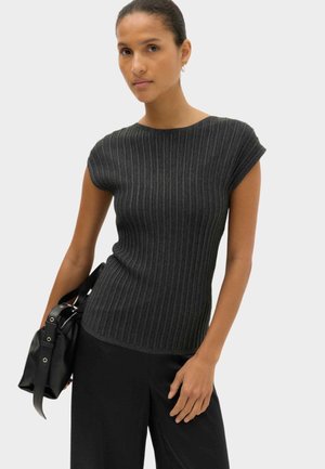 Femme portant un haut côtelé gris foncé à manches courtes et un pantalon noir, tenant un sac à main noir avec des boucles sur un fond uni.