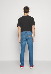 T-shirt scuro da uomo abbinato a jeans in denim azzurro chiaro. Presenta un classico design a cinque tasche e una patch in pelle sul retro. Texture liscia.