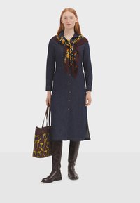 Vestido de mezclilla con botones, de manga larga y con aberturas laterales, combinado con una bufanda estampada y botas marrones hasta la rodilla, sosteniendo una bolsa de mano estampada.