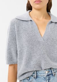 Siva pletenina iz volne z ovratnikom, kratkimi rokavi in globokim V-izreza, v kombinaciji z svetlo modrimi denim kratkimi hlačami. Mehka, teksturirana tkanina.