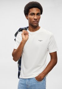 Baltas kokvilnas t-krekls ar īsām piedurknēm, uz kura krūtīm ir neliels melns logo. Gaiši zilas džinsu bikses un šūnu raksta krekls, kas pārklāts uz pleca.