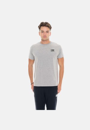 Jeune homme portant un t-shirt gris Leone et un pantalon noir, debout avec les bras détendus sur un fond blanc uni.
