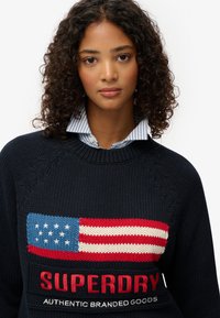 Granatowy sweter dziany z dużym wzorem amerykańskiej flagi z czerwonymi, białymi i niebieskimi akcentami oraz logo "SUPERDRY" poniżej.