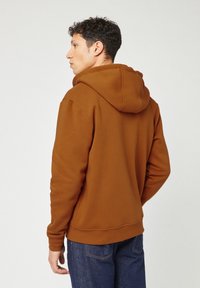 Giovane con capelli ricci e scuri, che indossa una felpa con cappuccio marrone e jeans blu, in piedi rivolto a sinistra con le mani rilassate lungo i fianchi.