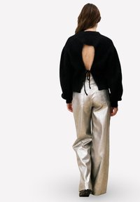 Zwarte wollen pullover met een uitsnijding aan de achterkant en een strikdetail, gecombineerd met zilverkleurige metallic wijde broek met hoge taille. Platte zwarte schoen zichtbaar.