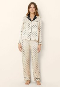 Beige polka dot pajama set made of smooth fabric, featuring long sleeves, black piping, and a chest pocket on the top. Pants have an elastic waist.