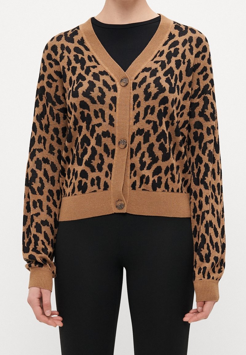 Cardigan boutonné imprimé léopard marron et noir avec poignets et ourlet côtelés, porté sur un haut noir et un pantalon noir.