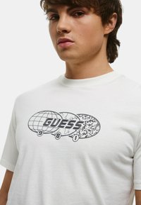 T-shirt bianco a maniche corte con stampa grafica che presenta la parola "GUESS" e design di globi in nero e grigio. Texture di cotone liscia.