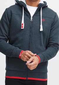 Sweat à capuche zippé gris foncé avec doublure blanche, accents rouges et cordons de serrage. Poignets et ourlet côtelés. Présente un logo sur la poitrine et une bande en bas.