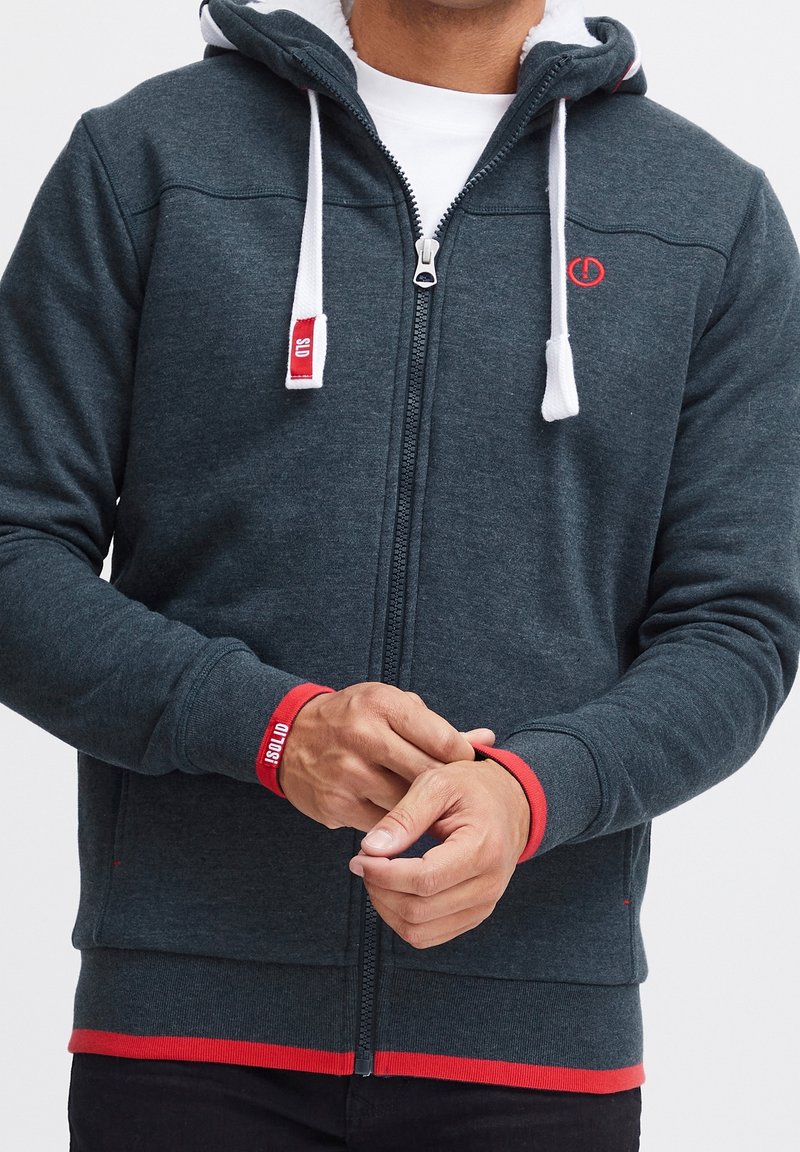 Sweat à capuche zippé gris foncé avec doublure blanche, accents rouges et cordons de serrage. Poignets et ourlet côtelés. Présente un logo sur la poitrine et une bande en bas.