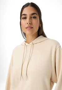Beige hoodie met een trekkoord, met een zachte textuur en een reliëflogo op de voorkant. Het ontwerp omvat een kangoeroezak.