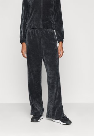 Persoon die losse zwarte velours sweatpants draagt met een bijpassend zip-up jack en zwart-witte sneakers tegen een effen achtergrond.