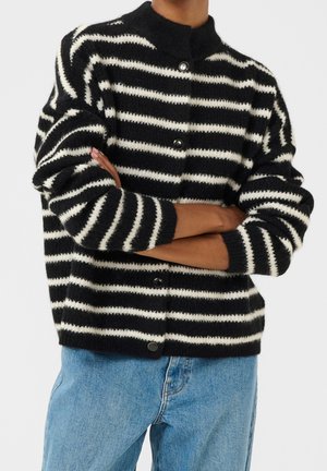Personne portant un pull en laine à rayures noires et blanches avec boutons, bras croisés, assorti à un jean en denim bleu clair.