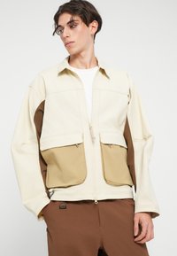 Svetle béžová bunda s hnědými akcenty, se zapínáním na zip vpředu, dvěma velkými kapsami a klasickým límcem. Hladká textura a neformální design.