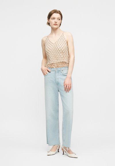 Γυναίκα που φοράει μπεζ αμάνικη μπλούζα από ανοιχτή πλέξη, ανοιχτό μπλε τζιν ψηλής μέσης με ξεφτισμένα τελειώματα και λευκά παπούτσια με τακούνι, στέκεται με το ένα χέρι στην τσέπη.