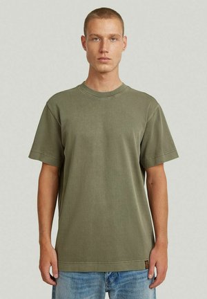 Giovane uomo con capelli corti che indossa una maglietta a girocollo verde oliva e jeans azzurri su uno sfondo semplice.