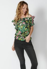 Top verde floreale senza spalline con accenti arricciati, caratterizzato da fiori rosa e bianchi su uno sfondo nero, abbinato a jeans neri.