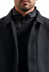 Zwarte wolmix jas met een omgeslagen kraag, gelaagd over een zwarte gewatteerde zip-up jack. Textiel met een gestructureerde stof en een gladde afwerking.