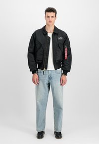 Mand med sort bomberjakke med lynlås og rød mærke, hvid skjorte, lyseblå jeans og sorte sko, der står foran en hvid baggrund.