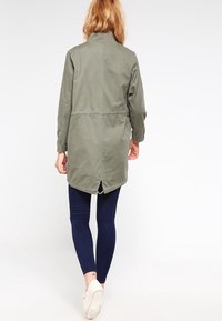 Persona con cabello largo y rubio, vistiendo una chaqueta verde oliva, leggings azul oscuro y zapatillas blancas, caminando alejándose sobre un fondo blanco.