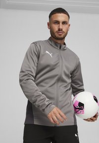 Grau zippered Jacke aus glattem Material mit hohem Kragen und weißem Logo; hält einen weißen und pinkfarbenen gemusterten Fußball.