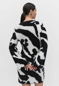 Neru valko-mustan kuviollisen neulemekon, jossa on pitkät liehuvat hihat, pehmeä pörröinen tekstuuri ja vartalonmyötäinen siluetti, nähtynä takaa.