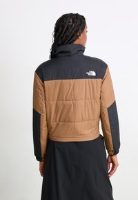 Apgriezta pūkainā jaka brūnā un melnā krāsā. Iezīme ar izšūtu rakstu, augstu apkakli un 'The North Face' logo aizmugurē. Elastīgas aproces.
