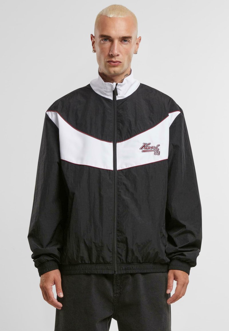 Veste noir et blanc avec un col haut, fermeture éclair et poignets élastiques. Caractéristiques du tissu texturé et logo brodé sur la poitrine.