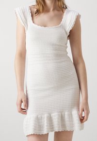 Robe blanche en tricot avec un décolleté carré, des manches ondulées bordées de dentelle, et un ourlet évasé à bords festonnés. Design en tissu texturé.