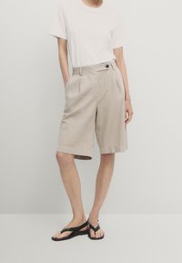 Beige bomuldsshorts med frontfolder, sidelommer og en knappedetalje i livet. Sammen med en ensfarvet hvid t-shirt og sorte sandaler.