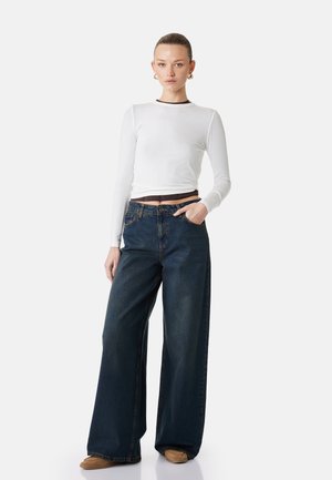Frau trägt weißes langärmliges Crop-Top, hoch taillierte, weit geschnittene dunkelblaue Jeans, braune Schuhe und goldene Creolen, steht vor weißem Hintergrund.