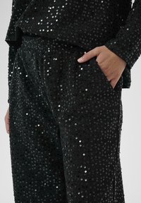 Černý outfit z flitrové látky s volným topem a kalhotami s elastickým pasem. Obsahuje boční kapsu, třpytivou texturu a kulaté flitry po celém povrchu.