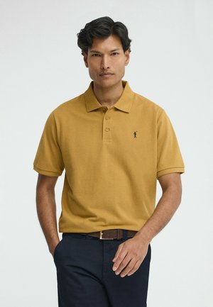 Uomo con polo giallo senape a maniche corte con logo e pantaloni blu scuro, in piedi con una mano in tasca su sfondo bianco.