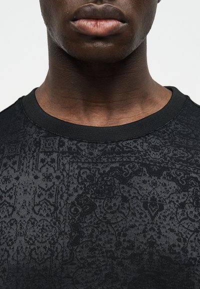 Κοντινό πλάνο του κατώτερου μέρους προσώπου και του λαιμού ενός ατόμου που φοράει μαύρο πουκάμισο με στρογγυλή λαιμόκοψη, με περίπλοκο σκοτεινό λουλουδένιο και γεωμετρικό σχέδιο.