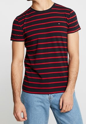T-shirt nero a maniche corte con strisce rosse orizzontali, caratterizzato da un piccolo logo sul petto, abbinato a jeans azzurri chiari.