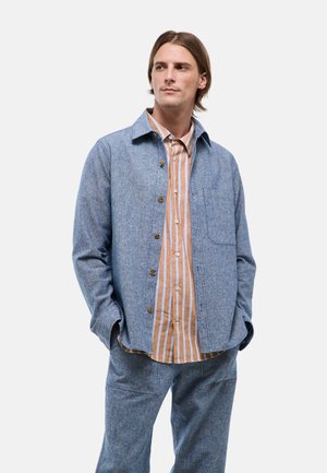 Homme aux cheveux mi-longs portant une veste bleue boutonnée sur une chemise rayée marron et blanche, les mains dans les poches, regardant sur le côté.