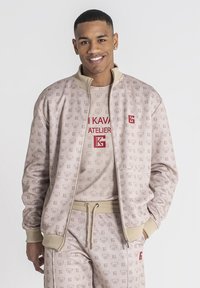 Beige bomberjack met herhalend logo-patroon, geribde manchetten en kraag. Gelaagd over een bijpassend T-shirt met rode tekst. Coördinerende joggingsbroek met trekkoord.