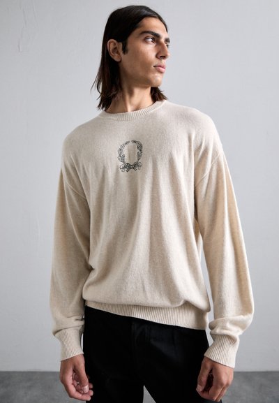 Ung man med axellångt mörkt hår som bär en beige tröja med ett svart dekorativt emblem och svarta byxor, som tittar åt sidan.