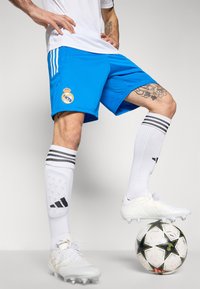 Blå atletikshorts med Real Madrid-logo, parret med hvidsokker med sorte striber og hvide fodboldstøvler, hviler på en sort og hvid stjerne-mønstrede bold.