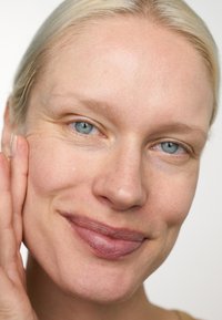 Primo piano di un volto con pelle chiara, capelli biondi, occhi azzurri e lentiggini sottili. Il soggetto tocca delicatamente la guancia, mettendo in risalto la bellezza naturale.