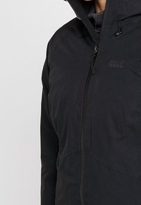 Svart huva jacka med dragkedja, tillverkad av slätt tyg. Har justerbara dragsko och subtilt logotypdetalj på bröstet.