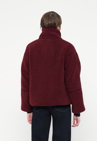 O persoană îmbrăcată într-o jachetă burgundă din fleece sherpa, cu guler înalt și mâneci lungi, stând cu fața întoarsă spre un fundal simplu.