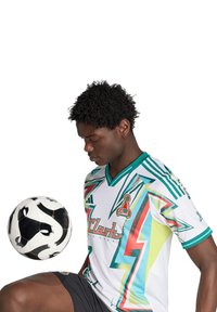 Junger männlicher Fußballspieler in buntem Trikot und schwarzen Shorts, der einen schwarz-weißen Fußball auf seinem Knie balanciert, vor einem weißen Hintergrund.