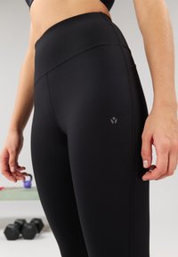 High-waisted black leggings with a smooth texture, discreet logo detail, and flat seams. Features a tapered fit and athletic design.
