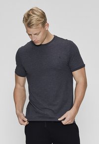 Lyshåret mand iført en tætsiddende mørkegrå T-shirt med rund hals, kigger nedad mens han justerer kanten af T-shirten med begge hænder.