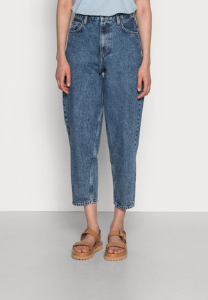 Persoon draagt blauwe, hooggetailleerde cropped jeans en tankleurige platform sandalen, staand tegen een effen witte muur en grijze vloer.