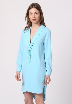 Joven mujer que lleva un vestido azul claro de manga larga con escote en V profundo y detalle de borlas, posando frente a un fondo claro y sencillo.