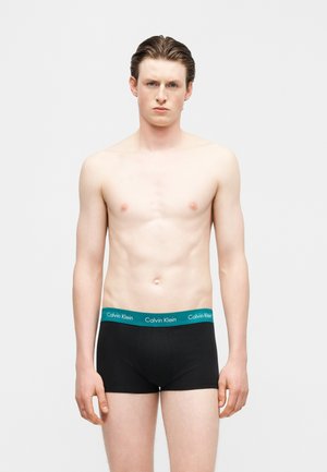 Νέος άνδρας με κοντά μαλλιά που φοράει μαύρα εσώρουχα Calvin Klein με μπλε ζωνάκι στη μέση, στέκεται μπροστά σε απλό φόντο.