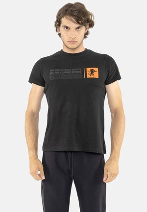 Mann trägt ein schwarzes T-Shirt mit dem Text "WE ARE COMBAT SPORTS" und einem orangefarbenen Löwenlogo auf der Brust, steht mit entspannt an den Seiten hängenden Armen.