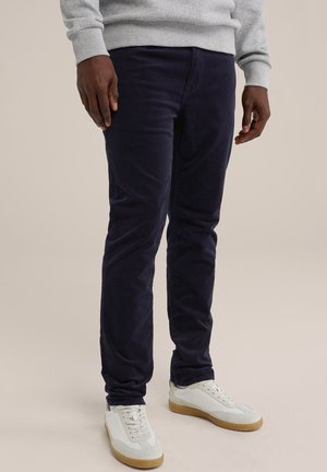 Mężczyzna w ciemnoniebieskich spodniach, szarym swetrze i białych trampkach z gumową podeszwą, stojący na jasnym, jednolitym tle.